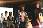 A fashion runway scene with models walking, showcasing diverse outfits in an indoor setting. The focus is on a shirtless male model wearing a colorful jacket, surrounded by other models displaying unique and stylish garments.