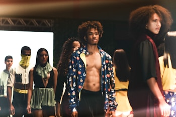 A fashion runway scene with models walking, showcasing diverse outfits in an indoor setting. The focus is on a shirtless male model wearing a colorful jacket, surrounded by other models displaying unique and stylish garments.