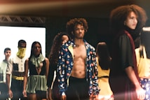 A fashion runway scene with models walking, showcasing diverse outfits in an indoor setting. The focus is on a shirtless male model wearing a colorful jacket, surrounded by other models displaying unique and stylish garments.