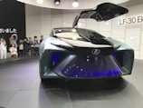 A futuristic concept car with a sleek design and gullwing doors open, showcasing advanced technology and stylish aesthetics. A group of people are observing and taking pictures, indicating a public exhibition or auto show setting.