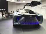 A futuristic concept car with a sleek design and gullwing doors open, showcasing advanced technology and stylish aesthetics. A group of people are observing and taking pictures, indicating a public exhibition or auto show setting.
