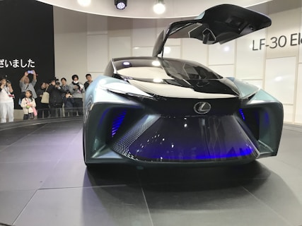 A futuristic concept car with a sleek design and gullwing doors open, showcasing advanced technology and stylish aesthetics. A group of people are observing and taking pictures, indicating a public exhibition or auto show setting.