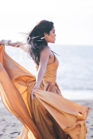 A woman stands gracefully on a sandy beach, with the ocean in the background. She is wearing an elegant, flowing orange gown that billows in the breeze. Her dark hair is swept back, and she wears a bracelet and earrings as accessories. The scene is bathed in soft, natural light, giving it a serene and ethereal quality.