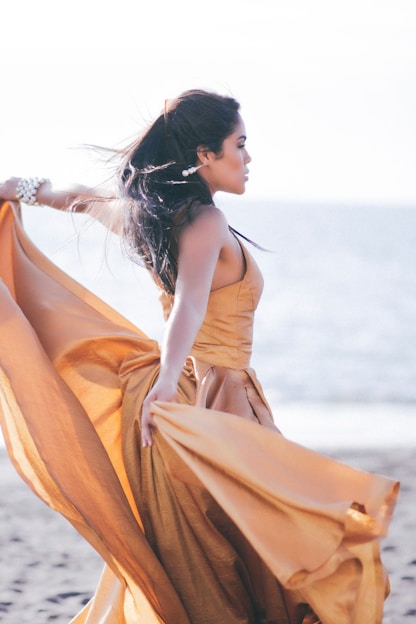 A woman stands gracefully on a sandy beach, with the ocean in the background. She is wearing an elegant, flowing orange gown that billows in the breeze. Her dark hair is swept back, and she wears a bracelet and earrings as accessories. The scene is bathed in soft, natural light, giving it a serene and ethereal quality.