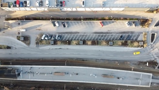 An aerial view of a parking lot next to a road and a railway platform. The parking lot contains scattered cars, while a few vehicles can be seen on the road. The railway platform is parallel to the road and appears to be quiet.