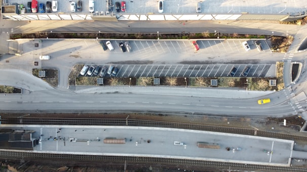 An aerial view of a parking lot next to a road and a railway platform. The parking lot contains scattered cars, while a few vehicles can be seen on the road. The railway platform is parallel to the road and appears to be quiet.
