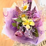 A vibrant floral arrangement is composed of various artificial flowers including yellow lilies, white daisies with yellow centers, and purple flora. The bouquet is wrapped in decorative purple and pink translucent wrapping paper, which adds a festive and delicate touch to the overall presentation.