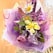 A vibrant floral arrangement is composed of various artificial flowers including yellow lilies, white daisies with yellow centers, and purple flora. The bouquet is wrapped in decorative purple and pink translucent wrapping paper, which adds a festive and delicate touch to the overall presentation.
