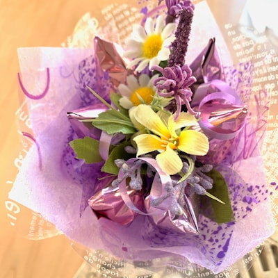 A vibrant floral arrangement is composed of various artificial flowers including yellow lilies, white daisies with yellow centers, and purple flora. The bouquet is wrapped in decorative purple and pink translucent wrapping paper, which adds a festive and delicate touch to the overall presentation.