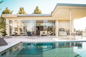 A modern, luxurious house with large glass windows opens onto a spacious patio with comfortable lounge chairs and an elegant outdoor dining set. The patio overlooks a serene swimming pool, reflecting the clear blue sky and surrounding greenery. The architecture features clean, minimalistic lines and a neutral color palette.