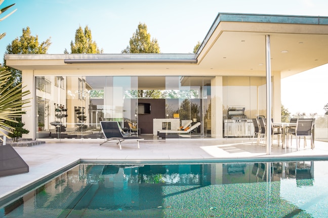 A modern, luxurious house with large glass windows opens onto a spacious patio with comfortable lounge chairs and an elegant outdoor dining set. The patio overlooks a serene swimming pool, reflecting the clear blue sky and surrounding greenery. The architecture features clean, minimalistic lines and a neutral color palette.