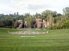A landscaped garden features a large grassy area with a round paved section in the center, bordered by manicured lawns. In the background, a series of cascading waterfalls flows over natural rock formations, surrounded by dense green trees and foliage.