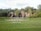 A landscaped garden features a large grassy area with a round paved section in the center, bordered by manicured lawns. In the background, a series of cascading waterfalls flows over natural rock formations, surrounded by dense green trees and foliage.