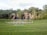 A landscaped garden features a large grassy area with a round paved section in the center, bordered by manicured lawns. In the background, a series of cascading waterfalls flows over natural rock formations, surrounded by dense green trees and foliage.