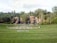 A landscaped garden features a large grassy area with a round paved section in the center, bordered by manicured lawns. In the background, a series of cascading waterfalls flows over natural rock formations, surrounded by dense green trees and foliage.