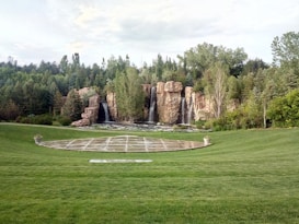 A landscaped garden features a large grassy area with a round paved section in the center, bordered by manicured lawns. In the background, a series of cascading waterfalls flows over natural rock formations, surrounded by dense green trees and foliage.