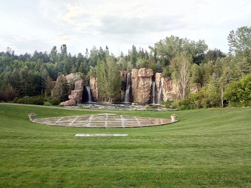 A landscaped garden features a large grassy area with a round paved section in the center, bordered by manicured lawns. In the background, a series of cascading waterfalls flows over natural rock formations, surrounded by dense green trees and foliage.