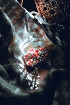 A glass jar filled with tiny origami stars in shades of red, white, and gray rests on a textured fabric. Sunlight filters through a wicker chair, casting intricate shadows and patterns across the scene, creating a harmonious blend of light and dark.