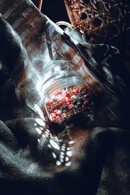 A glass jar filled with tiny origami stars in shades of red, white, and gray rests on a textured fabric. Sunlight filters through a wicker chair, casting intricate shadows and patterns across the scene, creating a harmonious blend of light and dark.