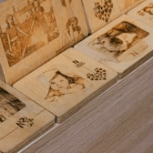 A collection of wooden plaques intricately engraved with images and text. The scenes include human figures, such as a child peeking from beneath a blanket, and what appears to be women holding hands, accompanied by motifs of hearts and text like 'N Seoul Tower'. The wood has a detailed finish and a warm hue.