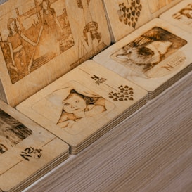 A collection of wooden plaques intricately engraved with images and text. The scenes include human figures, such as a child peeking from beneath a blanket, and what appears to be women holding hands, accompanied by motifs of hearts and text like 'N Seoul Tower'. The wood has a detailed finish and a warm hue.