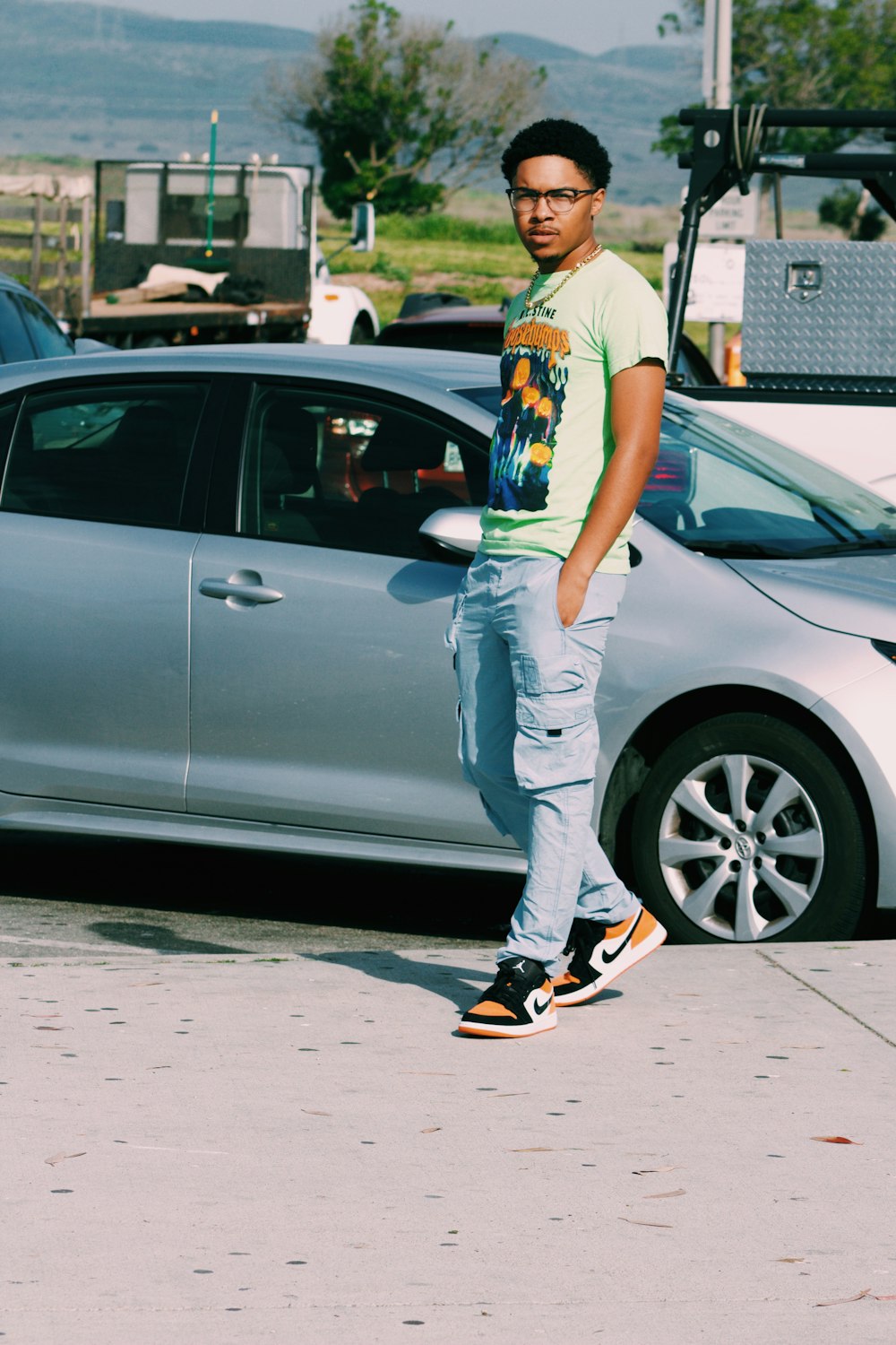 woman in white and yellow floral shirt and gray pants leaning on silver car