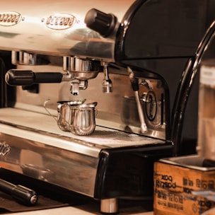 A shiny metal espresso machine with two small stainless steel milk pitchers placed on the drip tray, surrounded by dials and levers. The machine has a polished chrome surface with black handles and knobs, reflecting a warm light.