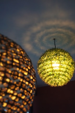 Two pendant lights with intricate designs hang from the ceiling. One has a greenish hue resembling foliage or leaves, while the other has a woven texture and emits a warm, golden light. The ceiling displays soft, swirling patterns from the light.