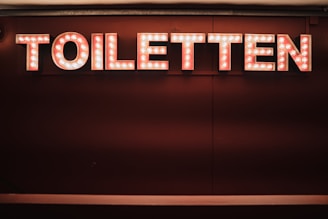 A sign displaying the word 'TOILETTEN' in bold, illuminated letters with a retro style. The background is a deep, rich shade, making the sign stand out prominently.