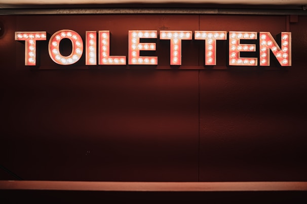 A sign displaying the word 'TOILETTEN' in bold, illuminated letters with a retro style. The background is a deep, rich shade, making the sign stand out prominently.