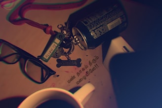 A collection of items on a surface includes a pair of black glasses, a keychain with the label 'ALLEN' and a dog tag reading 'SMITH,' a tipped over soda can, and some paper with text in another language. A mug with a beverage is also visible.
