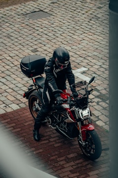 A person wearing full motorcycle gear, including a black helmet, is sitting on a red and black sports motorcycle parked on a cobblestone street. The image is taken from an elevated angle, capturing the rider and part of the patterned street.