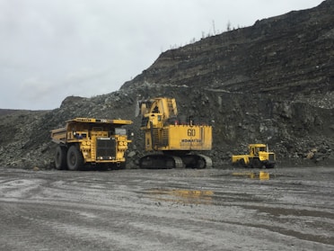 Heavy mining machinery is situated in a rocky terrain with piles of earth and rock in the background. There are three large vehicles, including a dump truck, a large excavator, and another piece of mining equipment.
