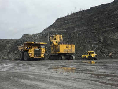 Heavy mining machinery is situated in a rocky terrain with piles of earth and rock in the background. There are three large vehicles, including a dump truck, a large excavator, and another piece of mining equipment.