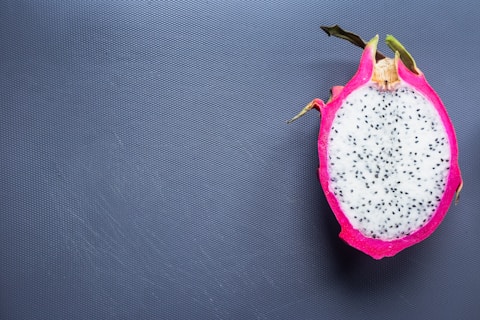 A sliced dragon fruit with vibrant pink skin and speckled white flesh lies on a textured gray surface, showcasing its unique appearance.