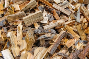 A pile of chopped firewood is stacked in a haphazard manner, showcasing various textures and grains. The pieces vary in size and shape, displaying a range of bark and exposed wood. Some logs feature darker bark, while others highlight lighter, freshly cut wood.