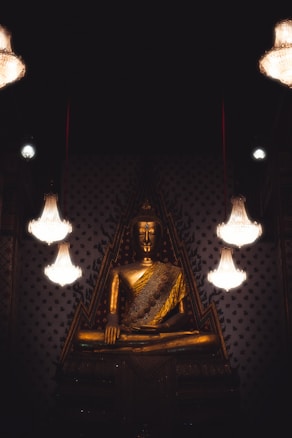 A large golden Buddha statue is prominently displayed, surrounded by ornate, intricately designed walls. Elegant chandeliers hanging from above cast a soft glow, enhancing the peaceful and reverent atmosphere of the scene.