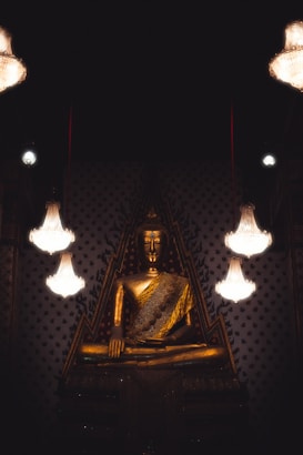 A large golden Buddha statue is prominently displayed, surrounded by ornate, intricately designed walls. Elegant chandeliers hanging from above cast a soft glow, enhancing the peaceful and reverent atmosphere of the scene.
