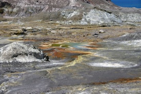 A barren landscape featuring rocky, uneven terrain with patches of mineral deposits. The surface is mostly gray and brown with some white crusts. A small, shallow pool with an orange tint is visible in the center, indicating geothermal activity.