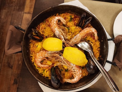 A pan of paella featuring large prawns and mussels arranged on top of the rice. Two lemon halves are placed in the center. The pan is accompanied by two spoons, and the dish is placed on a wooden table with a beige napkin.
