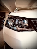 A close-up view of a white car's headlight, showcasing intricate details of the headlamp design with reflective elements. The car is parked indoors, and the environment is softly lit.