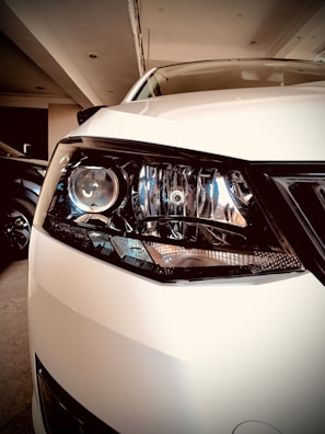 A close-up view of a white car's headlight, showcasing intricate details of the headlamp design with reflective elements. The car is parked indoors, and the environment is softly lit.