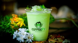 A green iced beverage in a plastic cup labeled 'Coffeefy' is surrounded by vibrant flowers and plants, creating a fresh and lively scene.