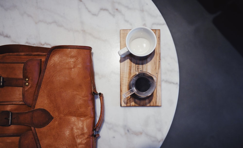 brown leather sling bag beside white ceramic mug on white round table