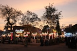 A bustling evening market scene with multiple food stalls illuminated by bright lights. The sky displays a warm, dusky tone as people gather around the stalls. 'Pista House Haleem' is prominently displayed on the signage, indicating a popular food item being served.