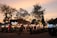 A bustling evening market scene with multiple food stalls illuminated by bright lights. The sky displays a warm, dusky tone as people gather around the stalls. 'Pista House Haleem' is prominently displayed on the signage, indicating a popular food item being served.