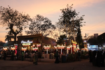A bustling evening market scene with multiple food stalls illuminated by bright lights. The sky displays a warm, dusky tone as people gather around the stalls. 'Pista House Haleem' is prominently displayed on the signage, indicating a popular food item being served.