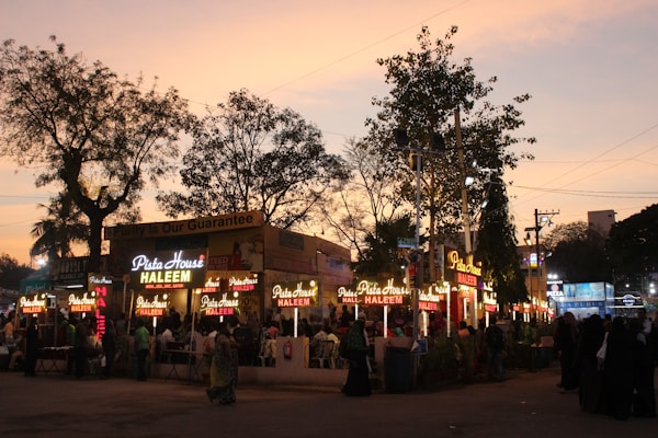 A bustling evening market scene with multiple food stalls illuminated by bright lights. The sky displays a warm, dusky tone as people gather around the stalls. 'Pista House Haleem' is prominently displayed on the signage, indicating a popular food item being served.