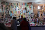 A market stall displaying a wide variety of colorful combs and hairbrushes. Numerous items hang on hooks and are carefully arranged on shelves. Two people are standing in front of the stall, seemingly interacting with a vendor. The scene is brightly lit by overhead lights, creating a vibrant atmosphere.
