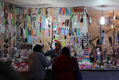 A market stall displaying a wide variety of colorful combs and hairbrushes. Numerous items hang on hooks and are carefully arranged on shelves. Two people are standing in front of the stall, seemingly interacting with a vendor. The scene is brightly lit by overhead lights, creating a vibrant atmosphere.